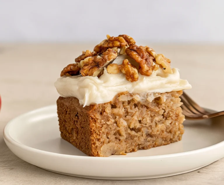 Slice of vegan applesauce cake topped with maple frosting and chopped walnuts on a white plate.